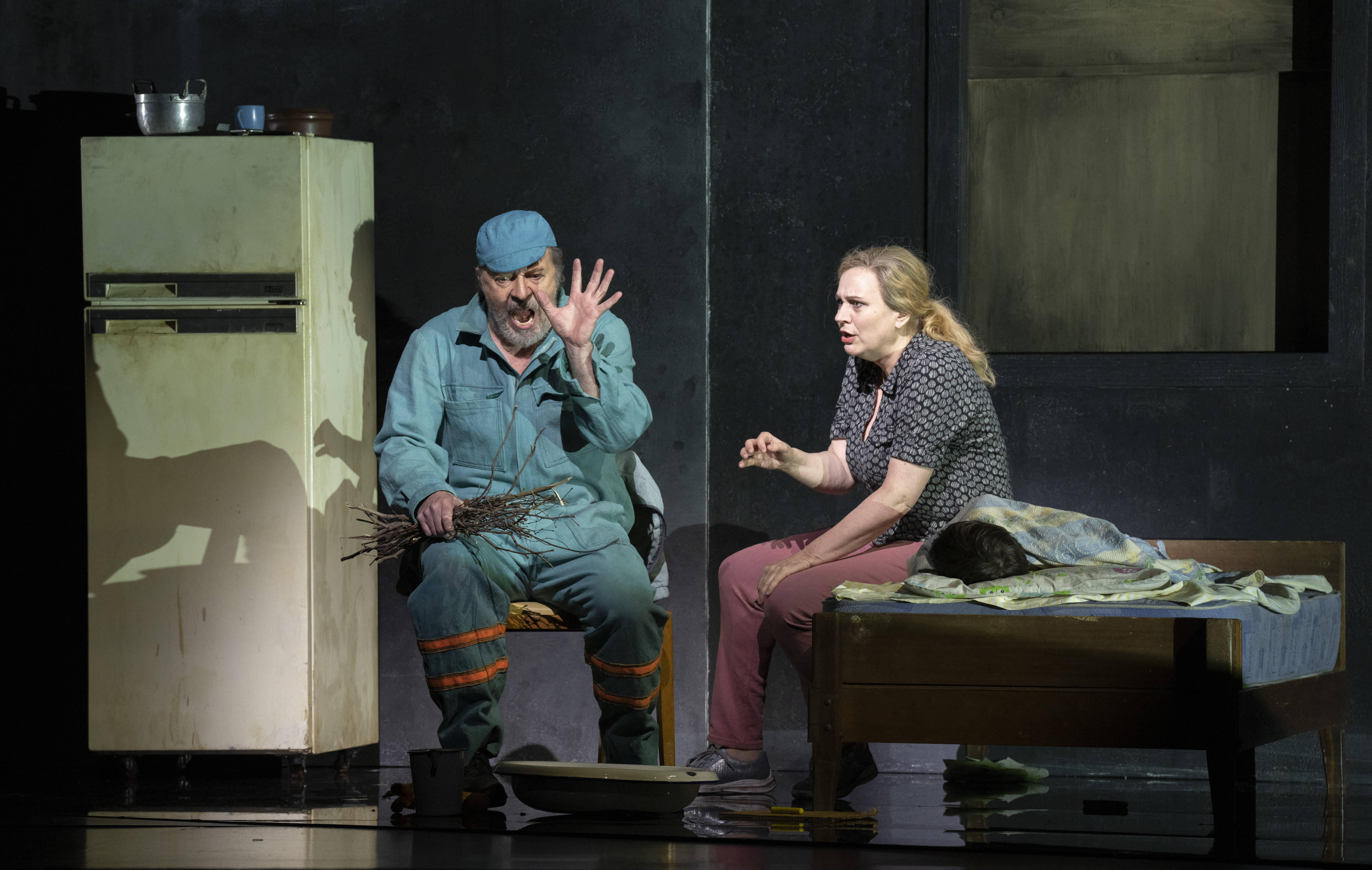 A man in blue work clothes and a woman in casual attire sit beside a bed in a dimly lit room; the man raises his hand, holding sticks, while the woman gestures emotionally. A fridge and kitchenware are in the background.