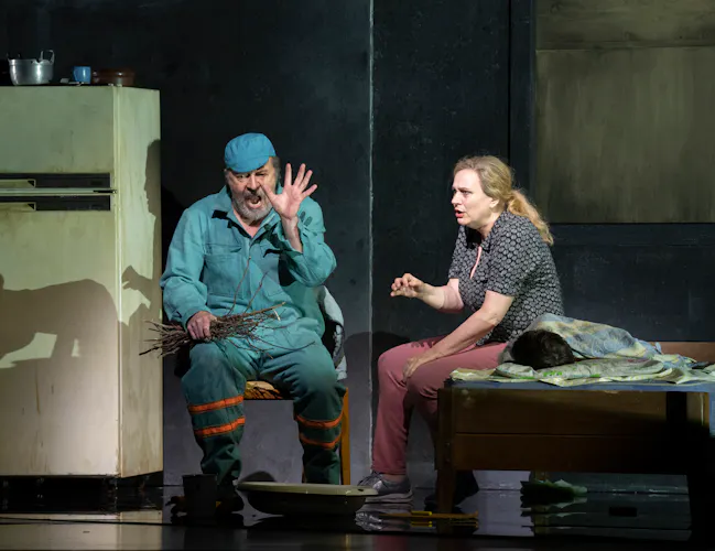 A man in blue work clothes and a woman in casual attire sit beside a bed in a dimly lit room; the man raises his hand, holding sticks, while the woman gestures emotionally. A fridge and kitchenware are in the background.