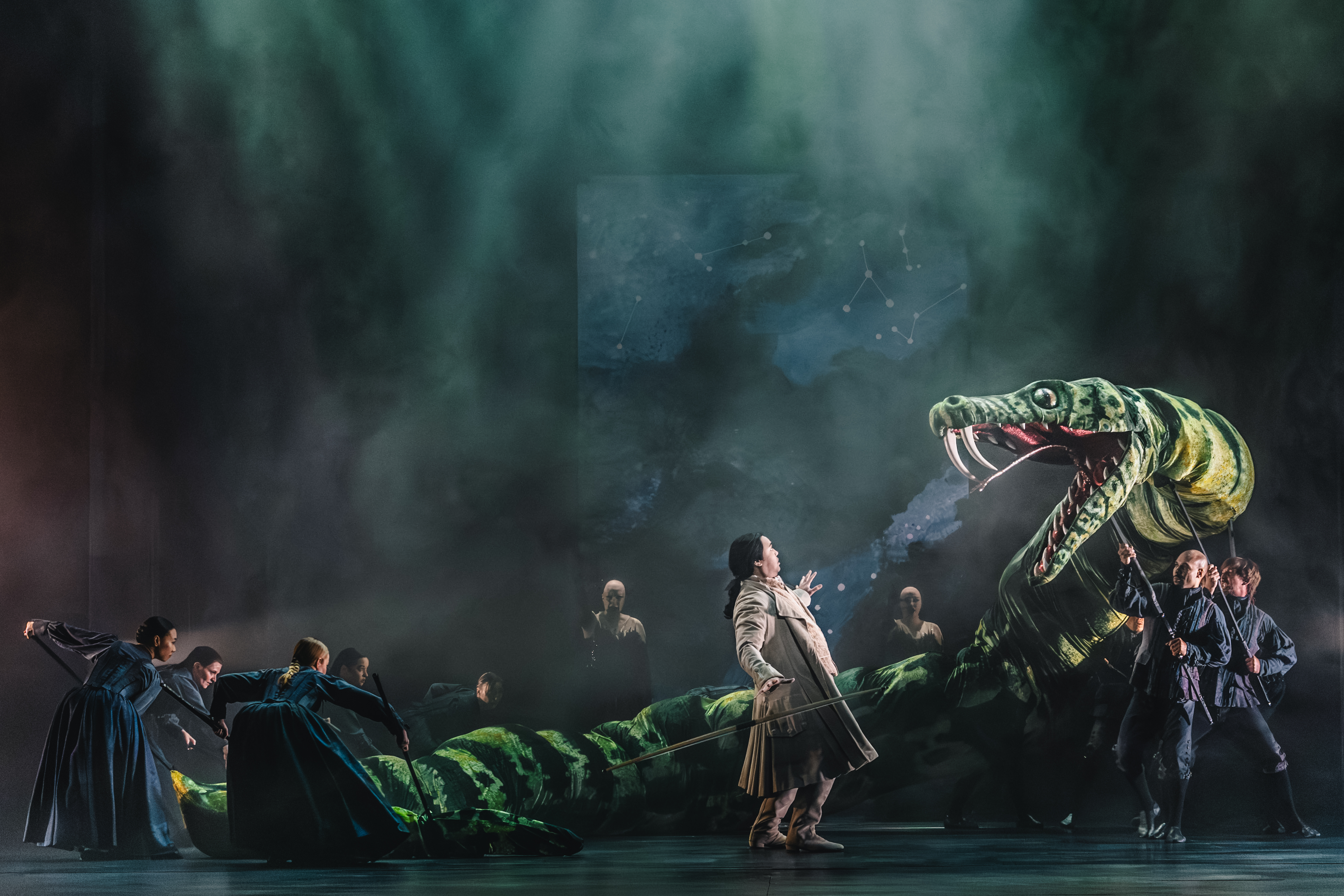 A giant green snake puppet with its mouth open, looms over an actor wearing a long brown coat.