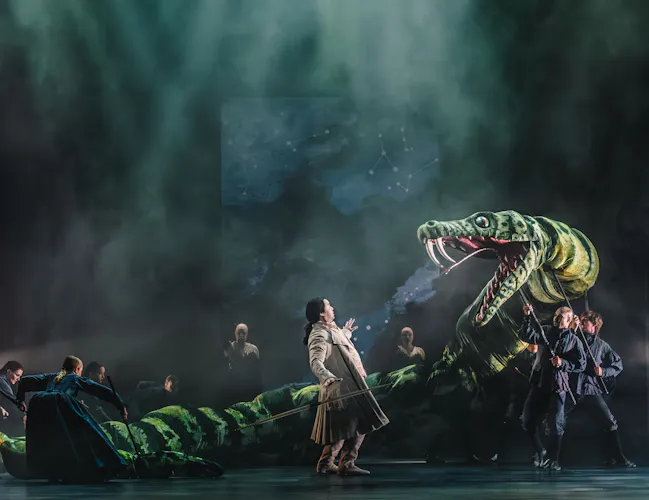 A giant green snake puppet with its mouth open, looms over an actor wearing a long brown coat.
