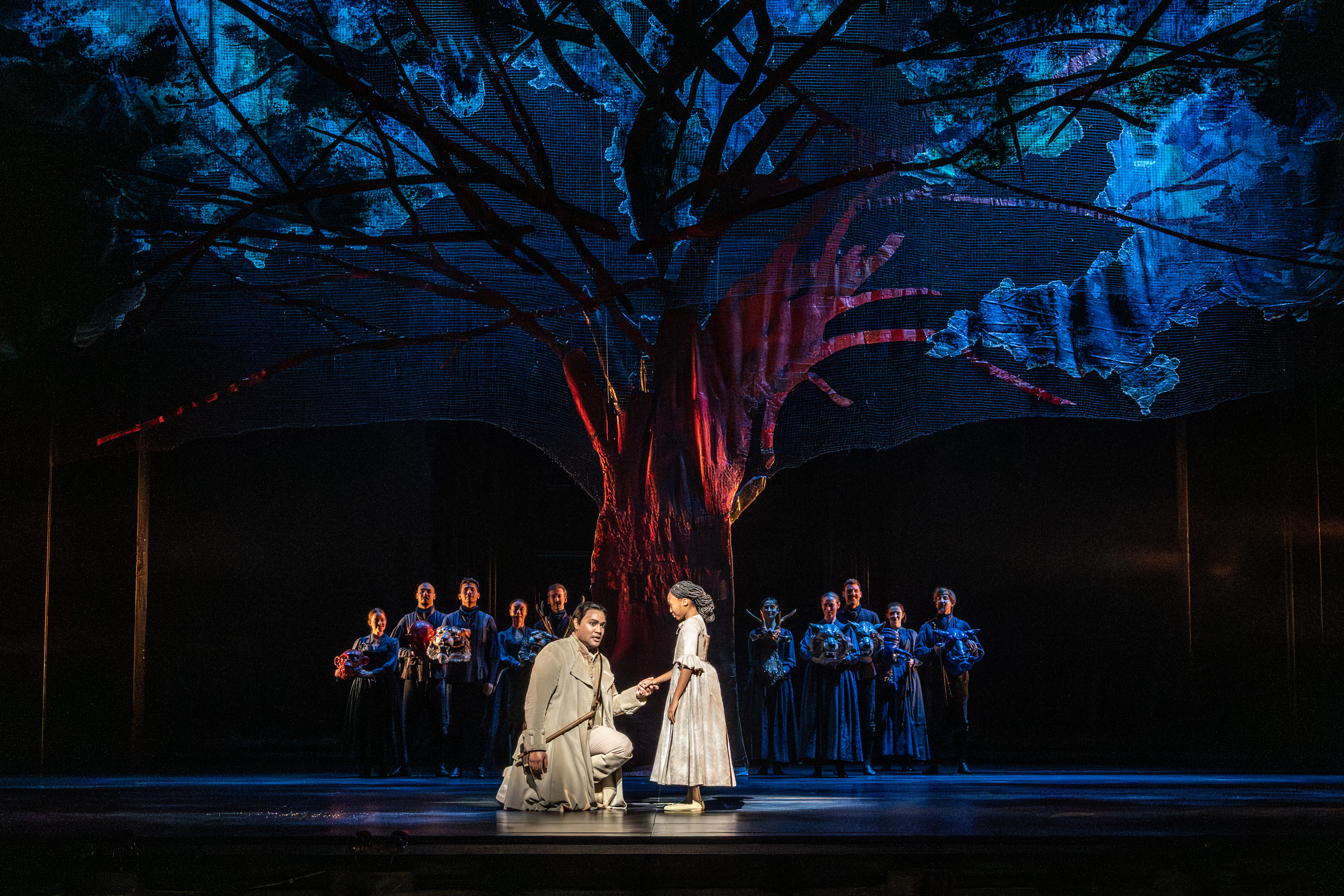 A man kneels holding hands with a young girl under a large, illuminated tree as people in dark clothing stand in the background,