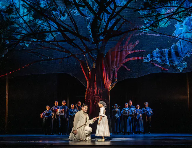 A man kneels holding hands with a young girl under a large, illuminated tree as people in dark clothing stand in the background,
