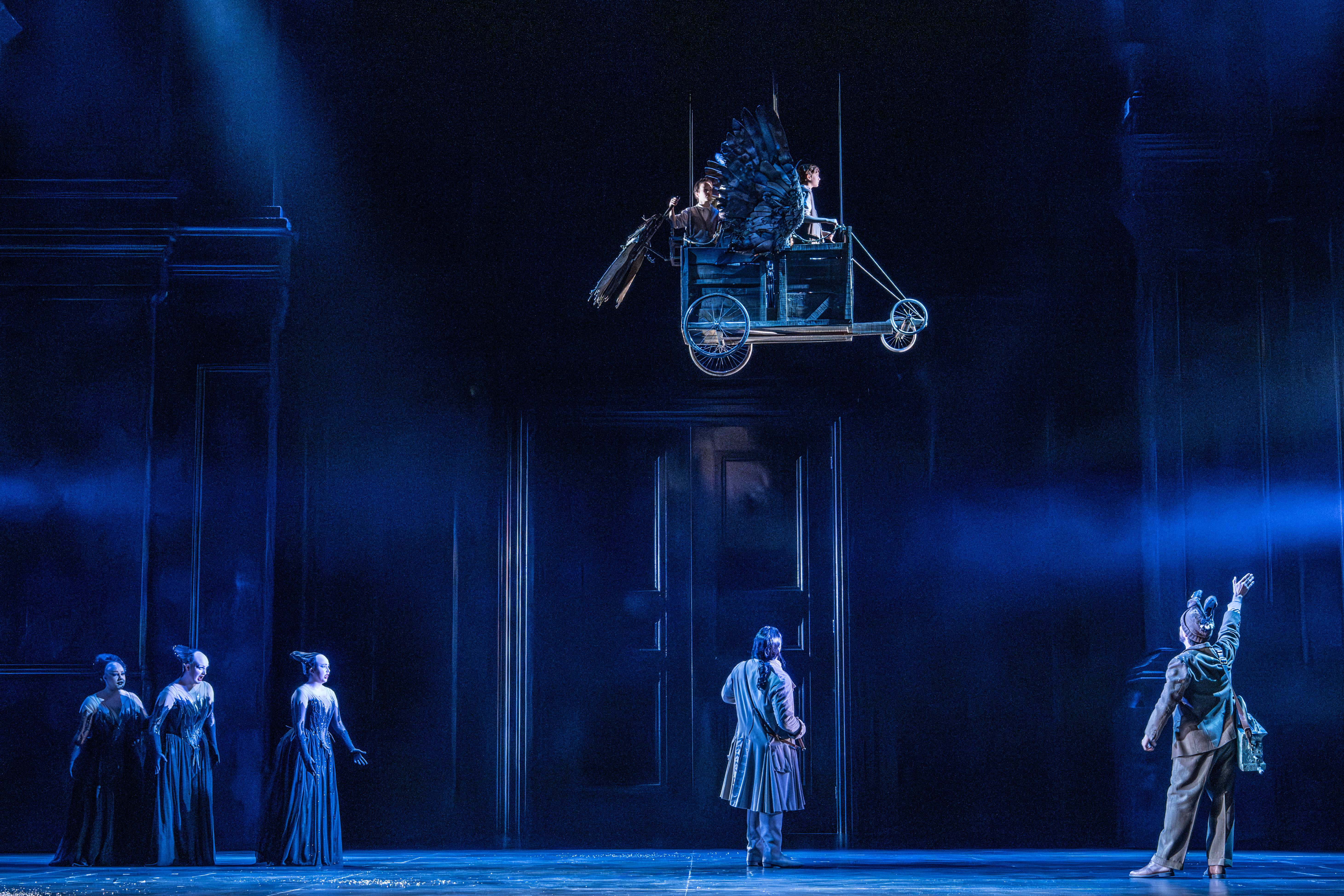 four actors on the ground in period costumes, looking up at a flying cart suspended above the stage. Three young performers sit in the cart as mechanical wings attached to it flap as the cart appears like it is flying across the stage. 