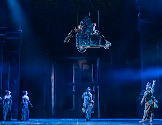 four actors on the ground in period costumes, looking up at a flying cart suspended above the stage. Three young performers sit in the cart as mechanical wings attached to it flap as the cart appears like it is flying across the stage.