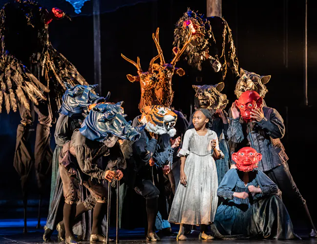 A girl in a light dress stands on stage surrounded by performers wearing large, elaborate animal masks and dark costumes.