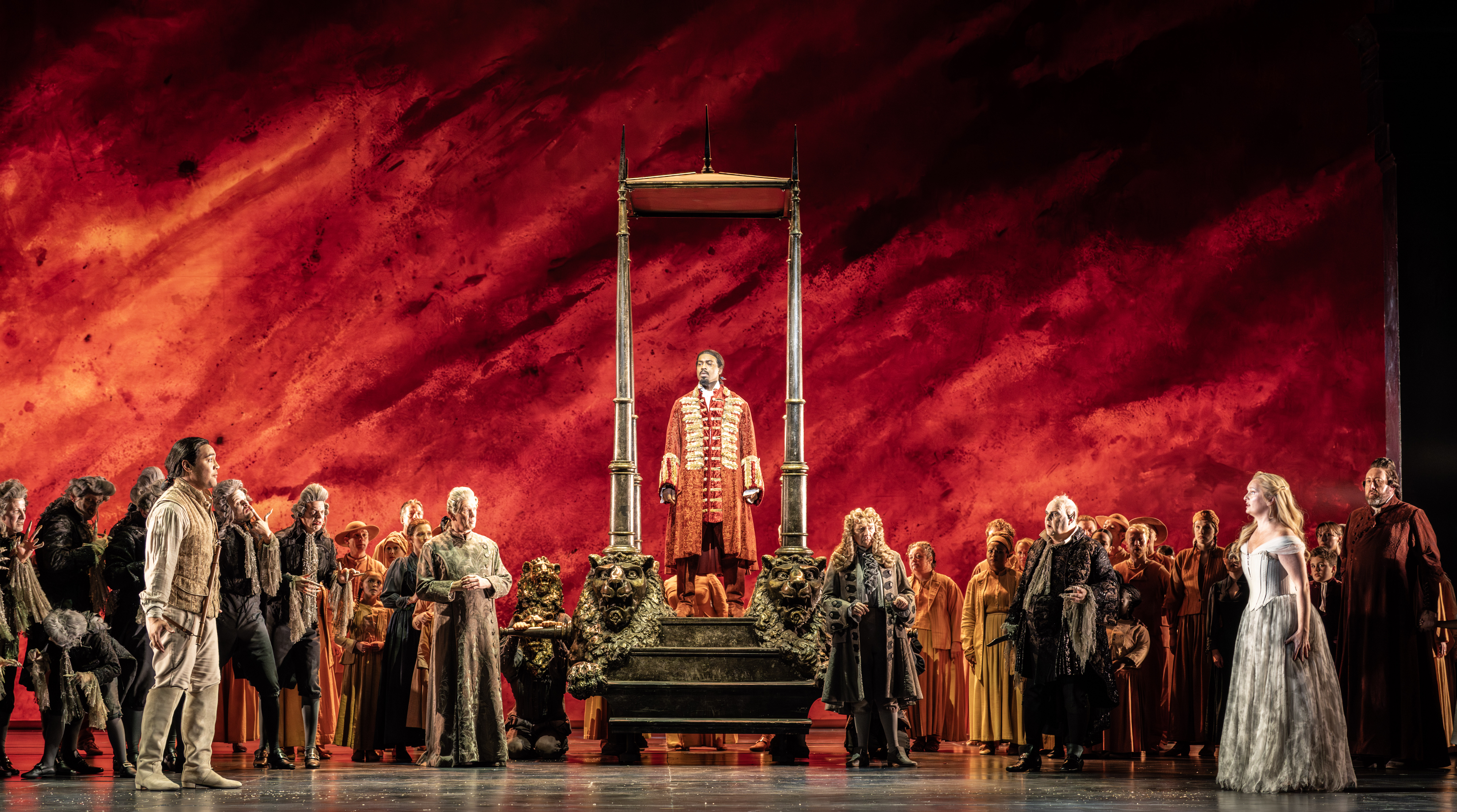 A man in ornate red frock coat stands on a raised platform resembling a guillotine, surrounded by people in period clothing,
