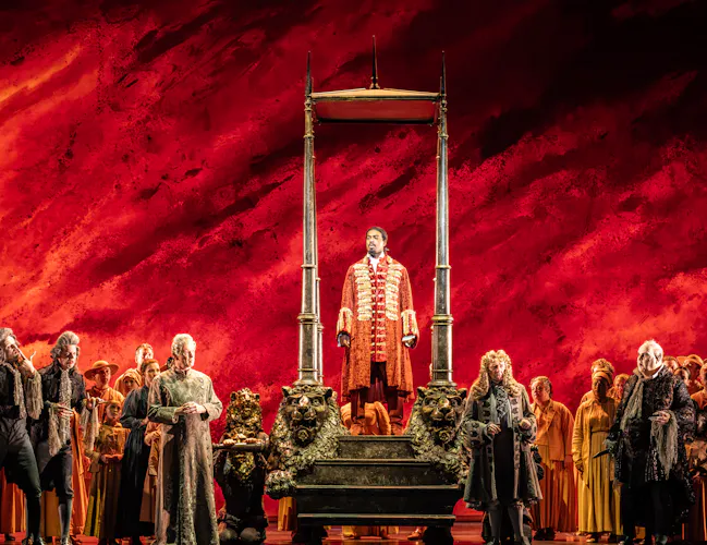 A man in ornate red frock coat stands on a raised platform resembling a guillotine, surrounded by people in period clothing,
