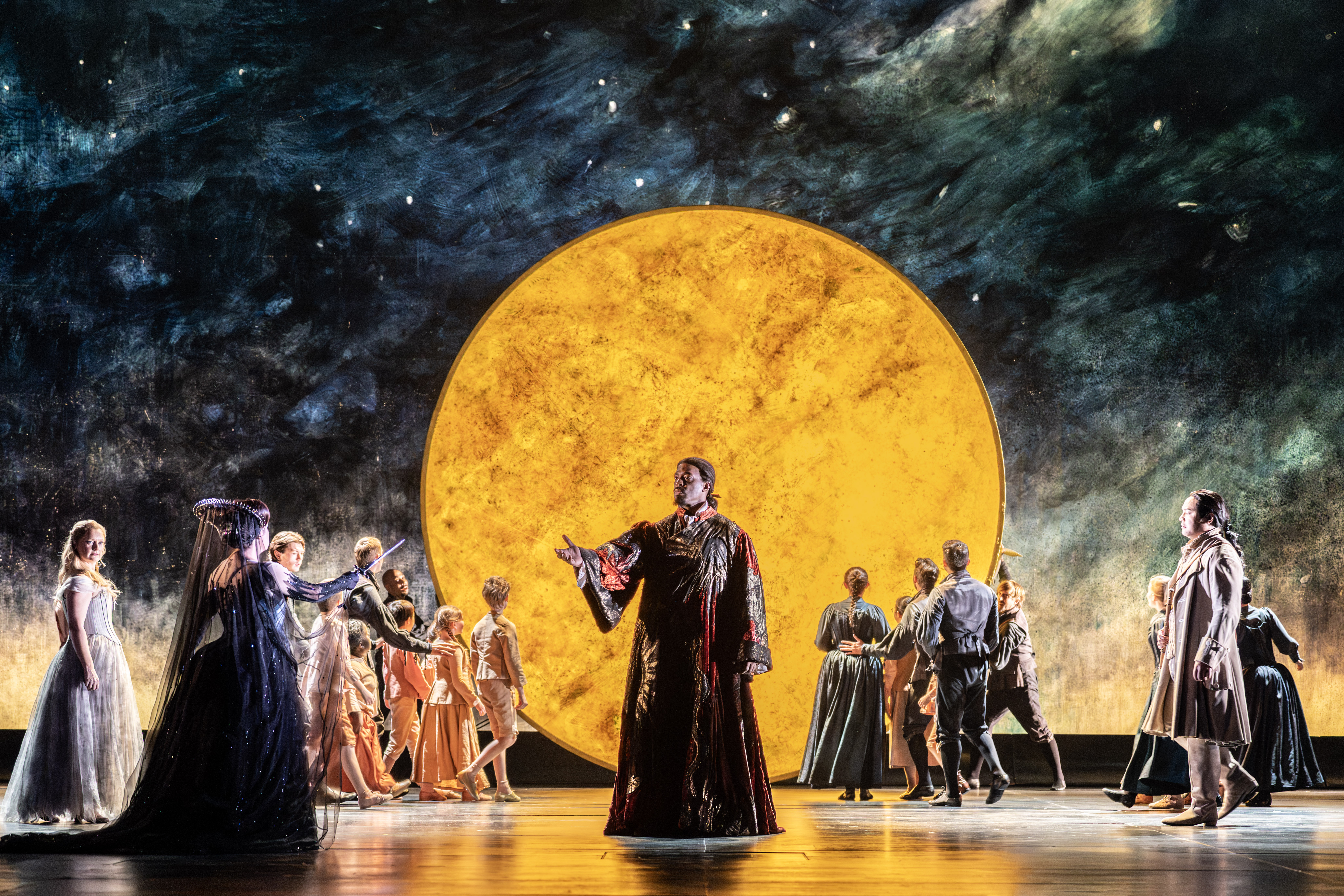 Performers in elaborate costumes stand on stage in front of a dramatic, large yellow circle resembling the moon, with a painted night sky backdrop. A man in a long red and gold cloak stands in the centre of the stage stretching out his hand towards a woman dressed in black. 