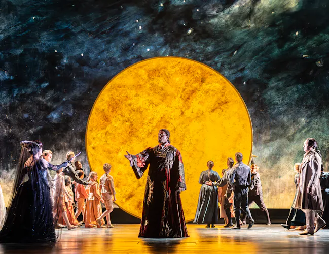 Performers in elaborate costumes stand on stage in front of a dramatic, large yellow circle resembling the moon, with a painted night sky backdrop. A man in a long red and gold cloak stands in the centre of the stage stretching out his hand towards a woman dressed in black.