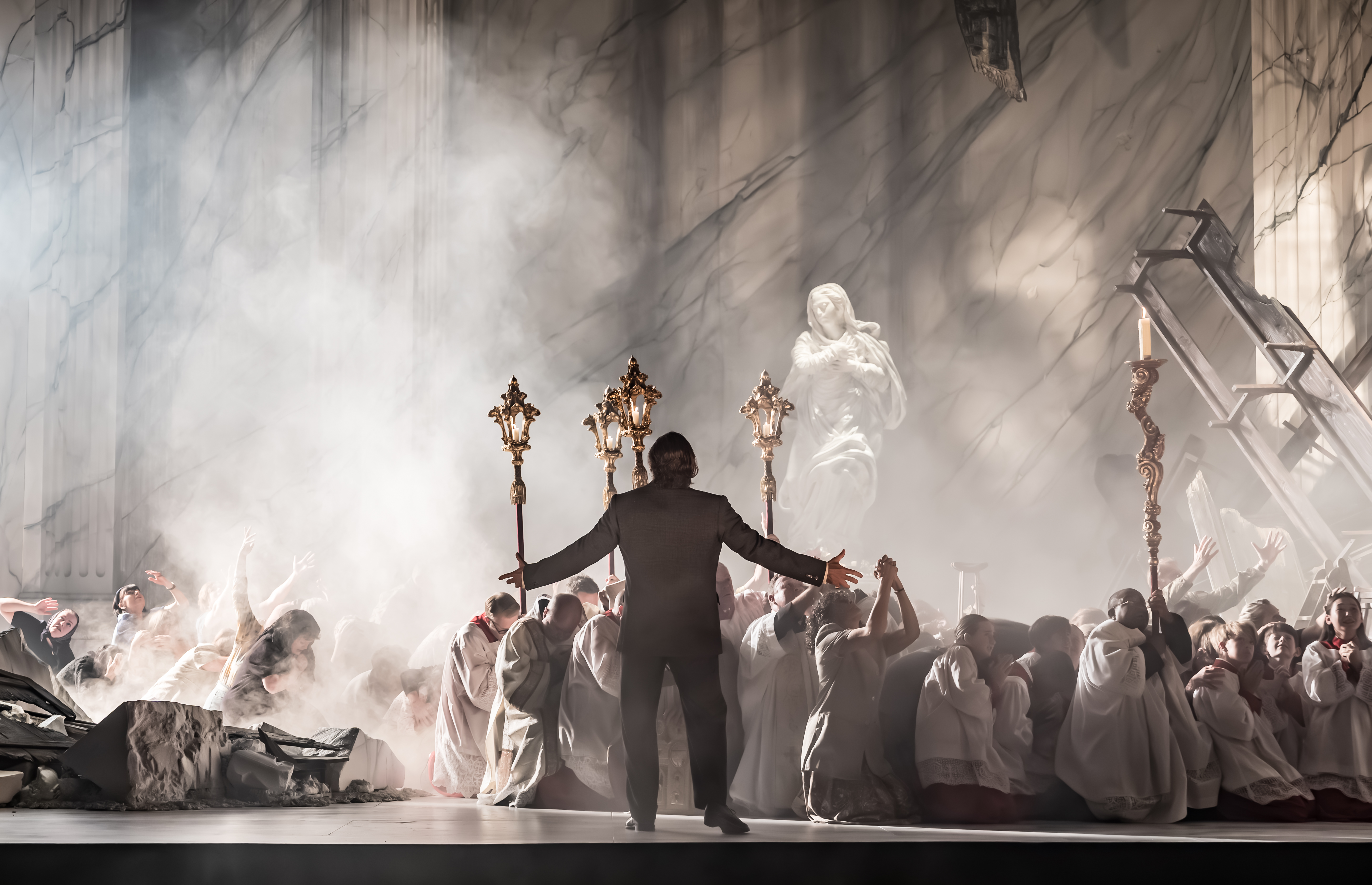 A man in a suit stands with arms outstretched before a group of kneeling people in white robes, holding ornate staffs, on a smoky stage with a marble statue and dramatic lighting.