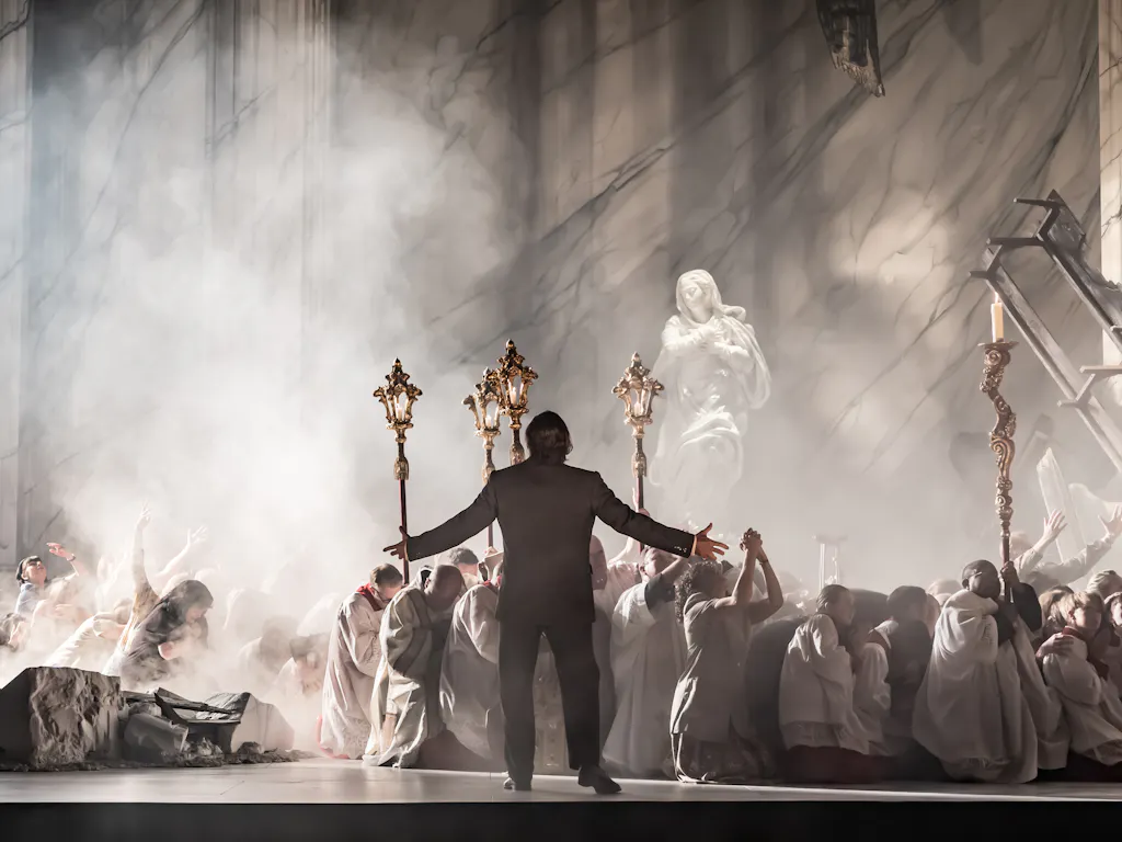 A man in a suit stands with arms outstretched before a group of kneeling people in white robes, holding ornate staffs, on a smoky stage with a marble statue and dramatic lighting.