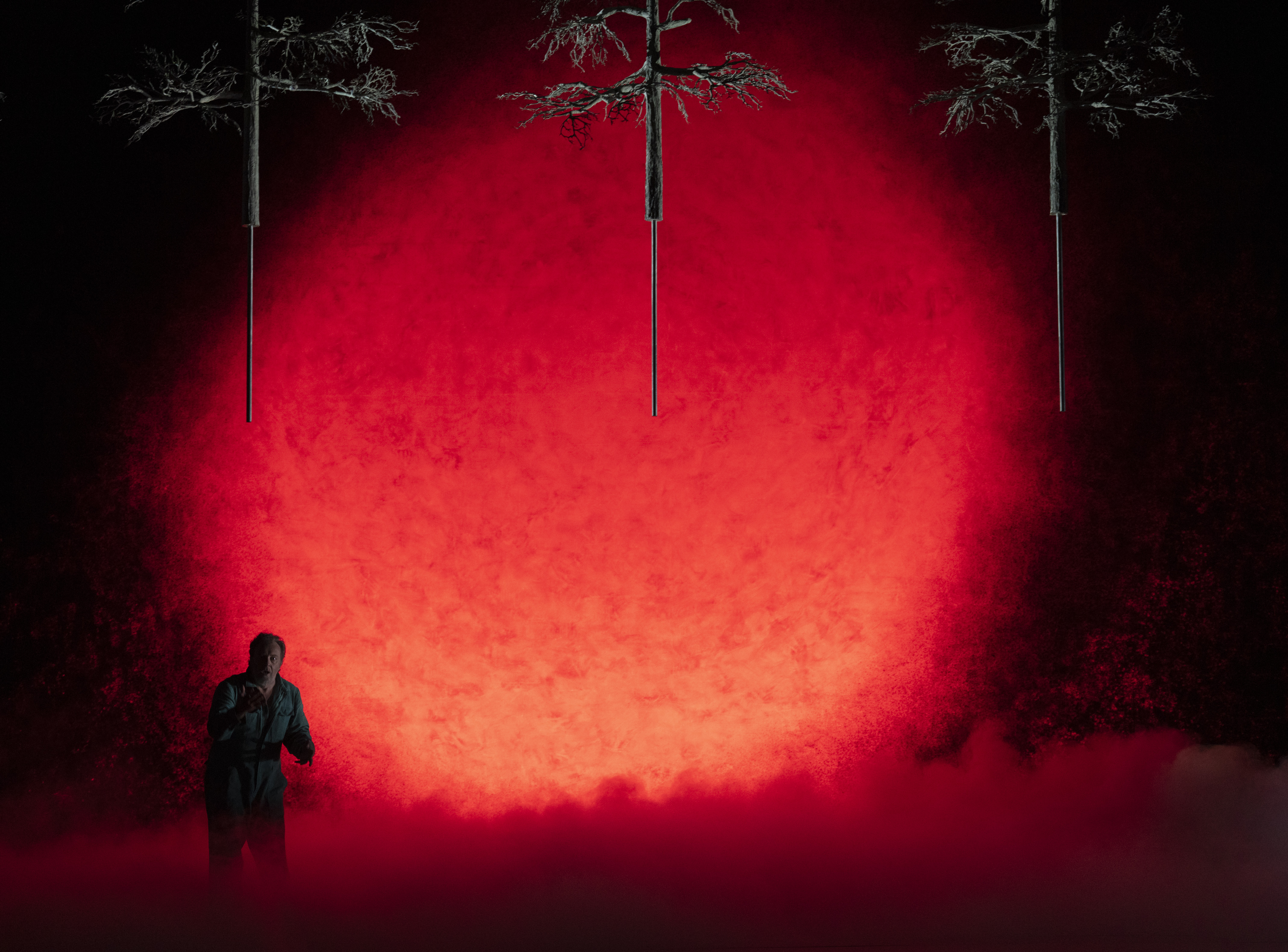 A person stands in a dark, smoky scene with a dramatic red circular light behind them. Three tree silhouettes hang upside down above him.