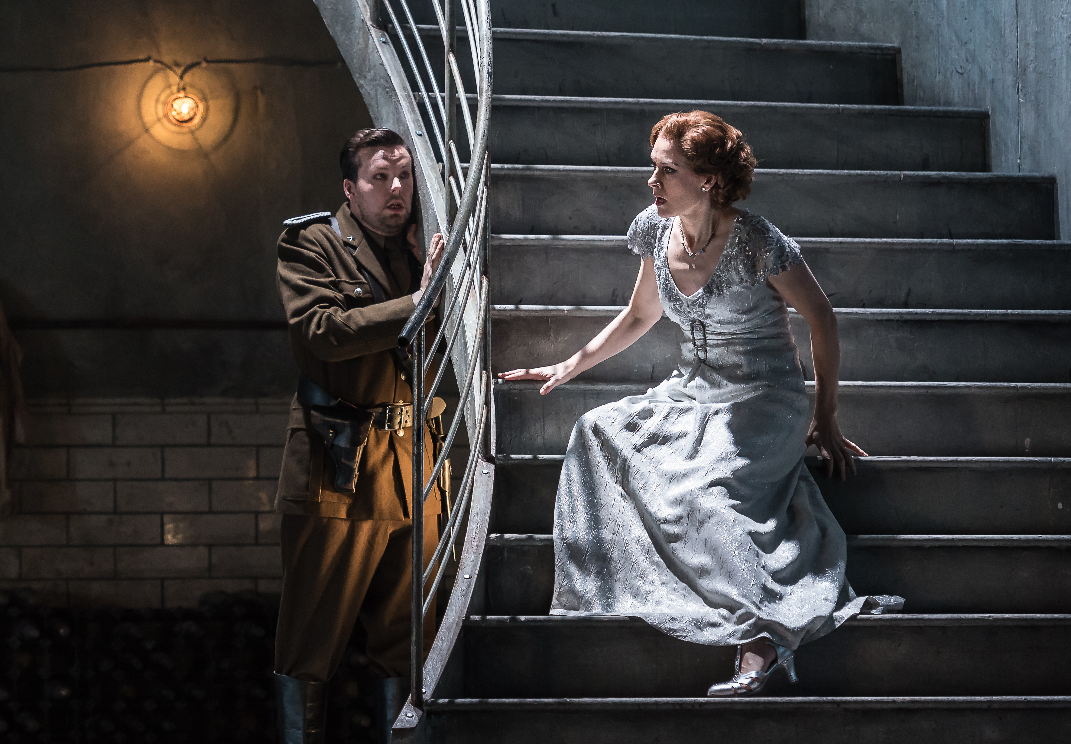 	 A man in a military uniform stands by a staircase railing, looking at a woman in a sparkling white gown who is sitting on a set of stairs. 