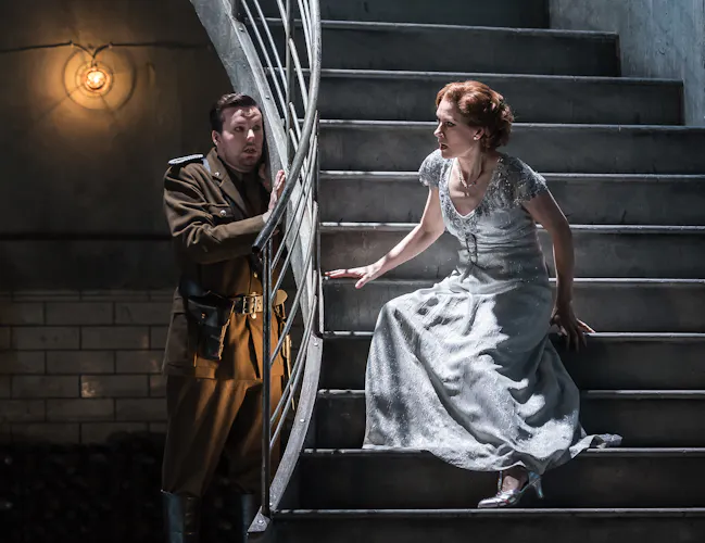 A man in a military uniform stands by a staircase railing, looking at a woman in a sparkling white gown who is sitting on a set of stairs.