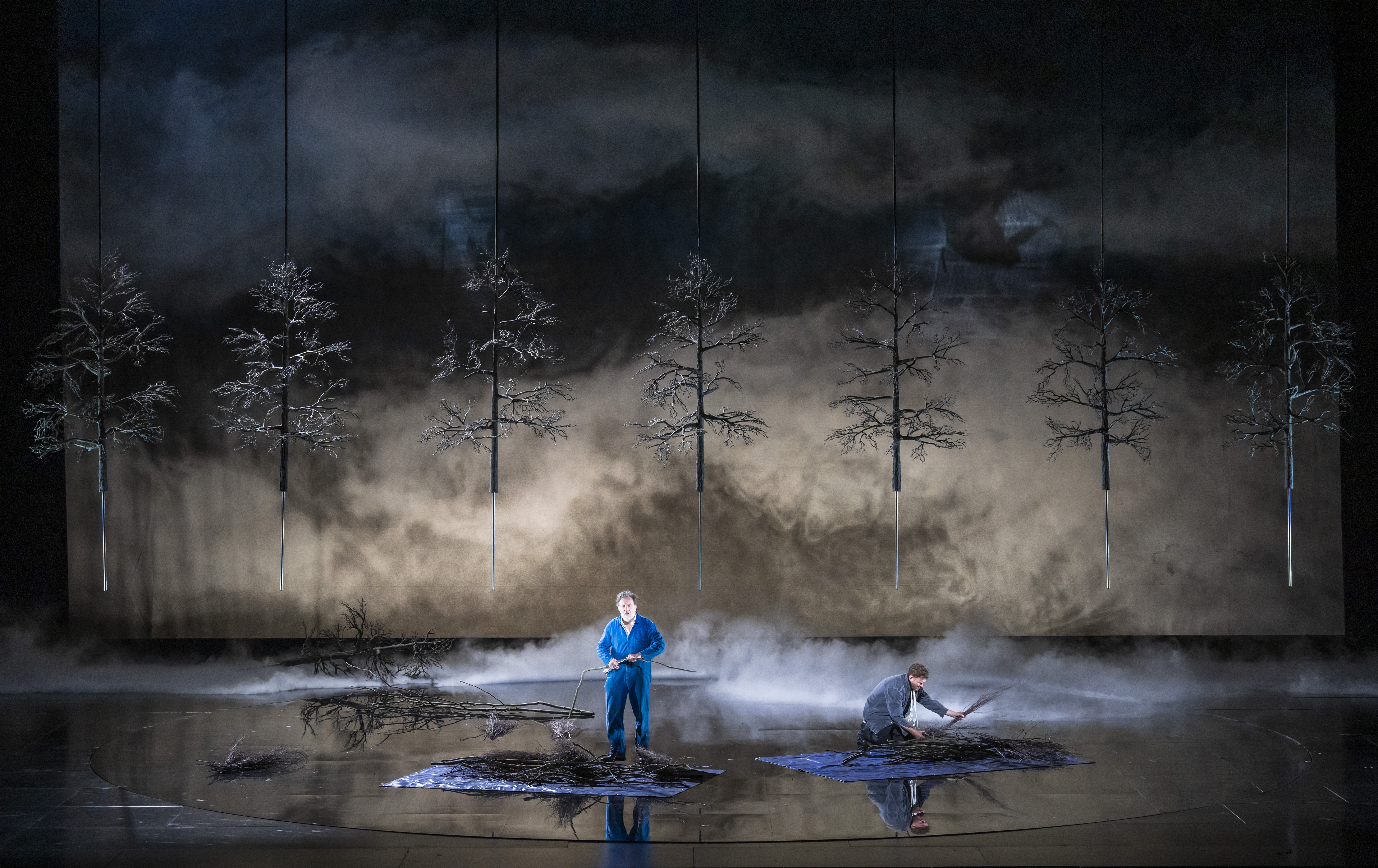 Two performers stand on a dimly lit stage with artificial trees and branches, surrounded by fog. One person stands holding a branch, while the other kneels, working with twigs.