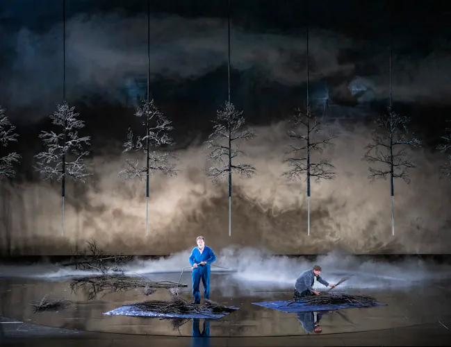 Two performers stand on a dimly lit stage with artificial trees and branches, surrounded by fog. One person stands holding a branch, while the other kneels, working with twigs.