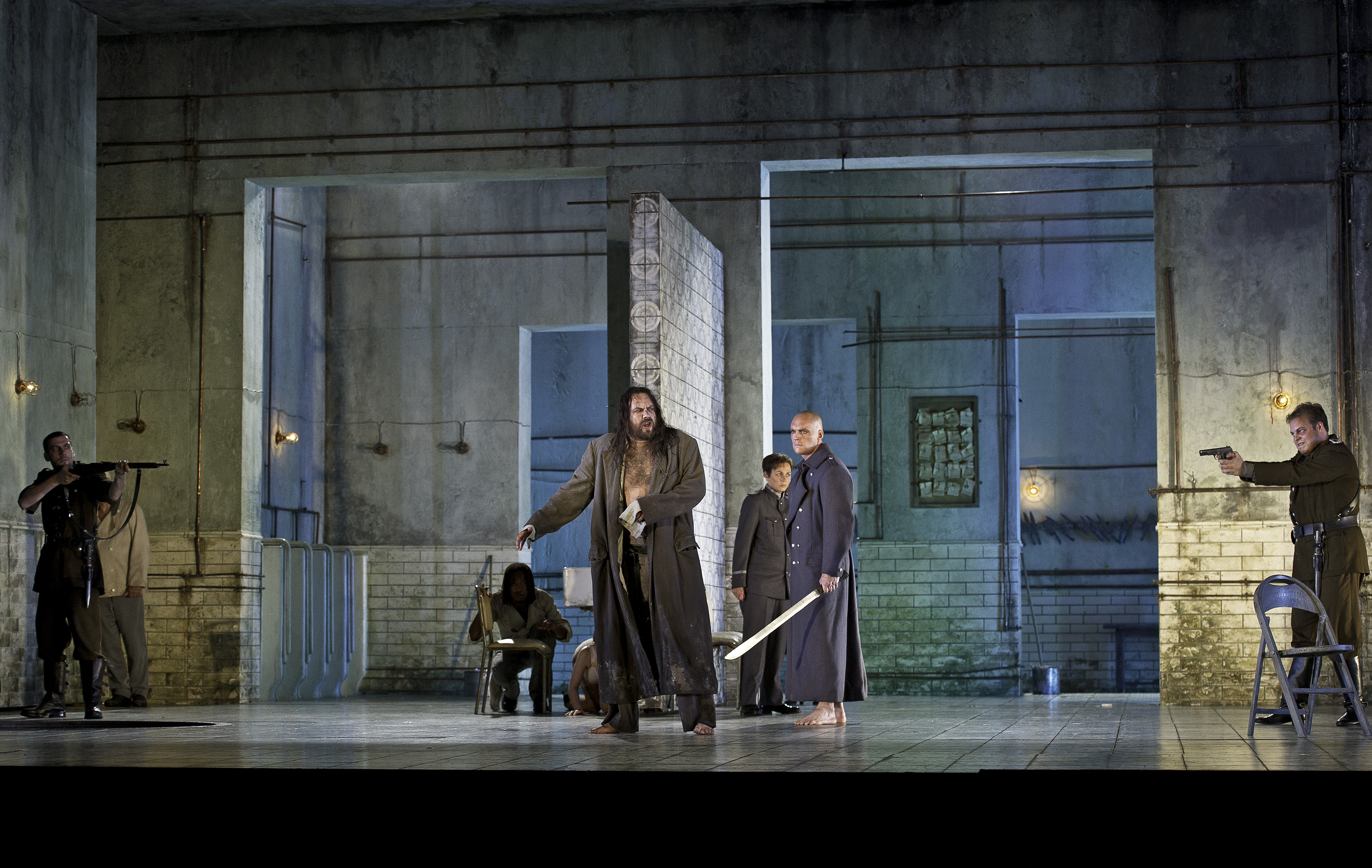 A man in a long brown coat stands at the centre of the room. Men in military uniform hold swords.  The set resembles a stark, industrial room with tiled walls and exposed pipes, lit with blue-toned lighting.