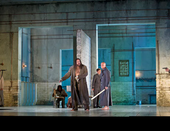 A man in a long brown coat stands at the centre of the room. Men in military uniform hold swords. The set resembles a stark, industrial room with tiled walls and exposed pipes, lit with blue-toned lighting.