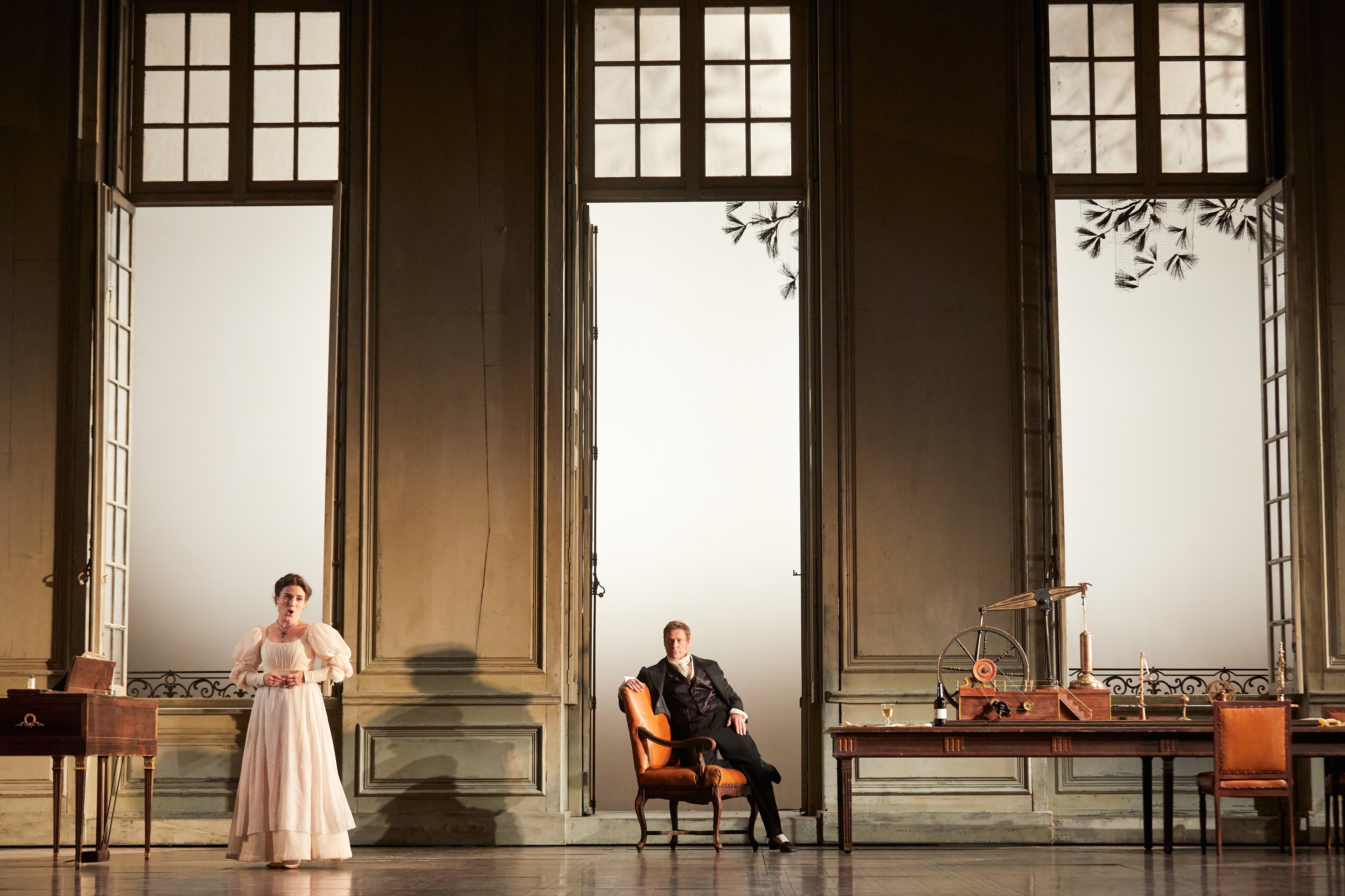 A woman in a white dress stands with her back to a man who is sat on an orange armchair and watching her. 