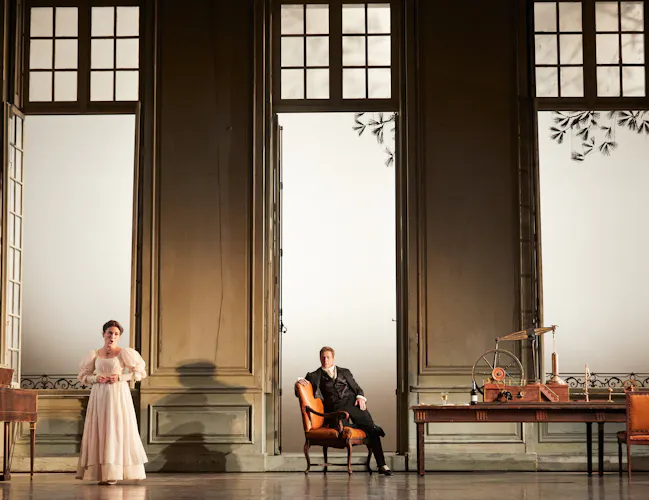 A woman in a white dress stands with her back to a man who is sat on an orange armchair and watching her.