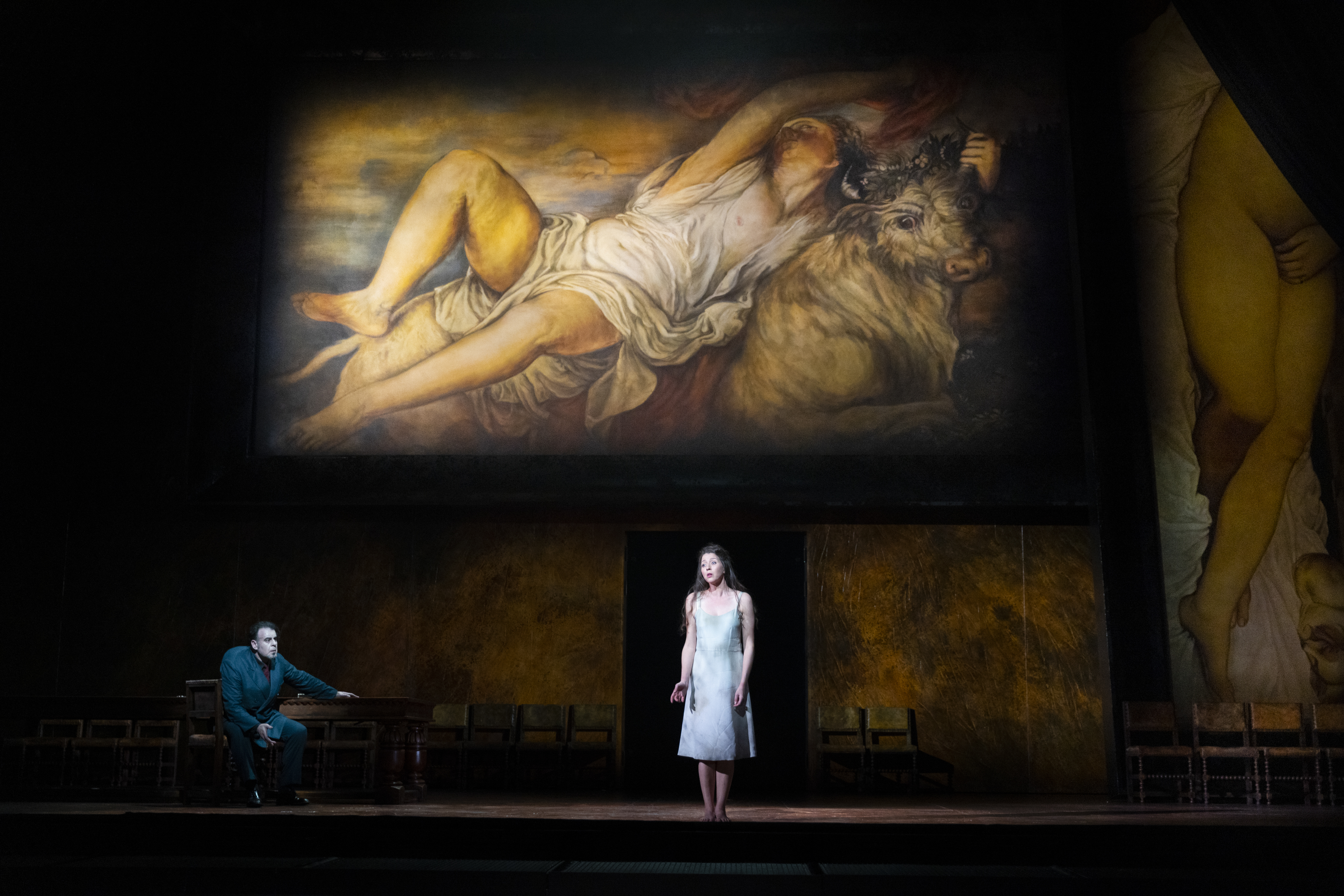 A woman in a short white dress stands centre stage. A painting is projected behind her. 