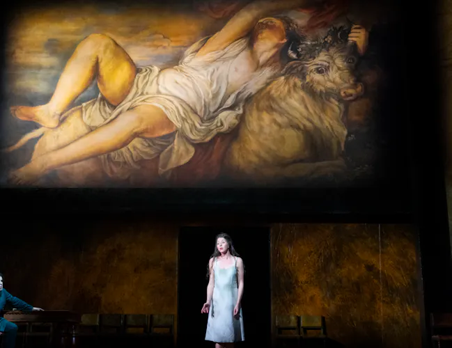 A woman in a short white dress stands centre stage. A painting is projected behind her.
