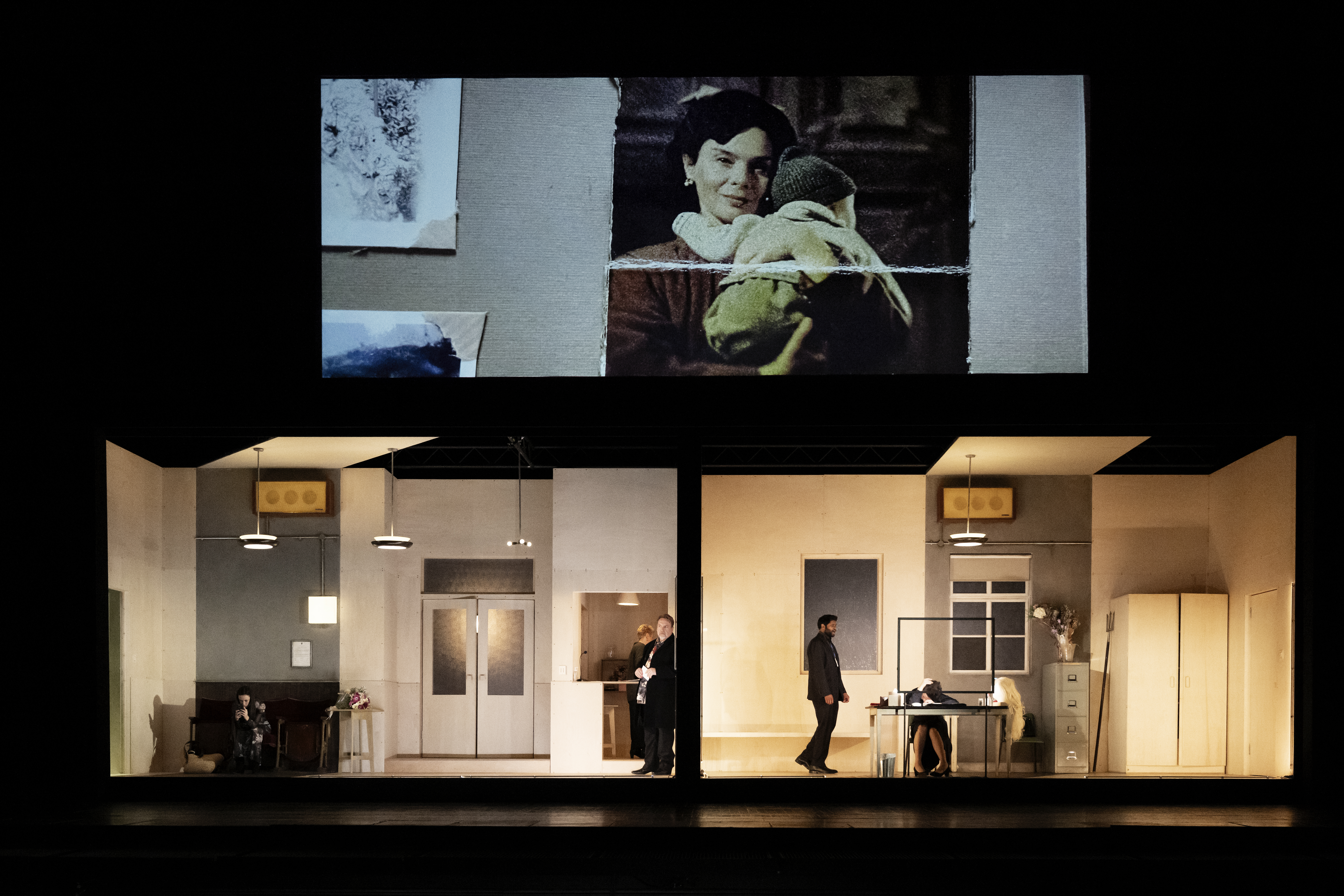 The stage is split into two rooms. Above the rooms is a screen. On the screen is a photo of a dark haired woman smiling and holding a toddler to her chest. . 