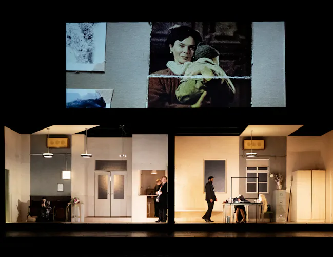 The stage is split into two rooms. Above the rooms is a screen. On the screen is a photo of a dark haired woman smiling and holding a toddler to her chest. .