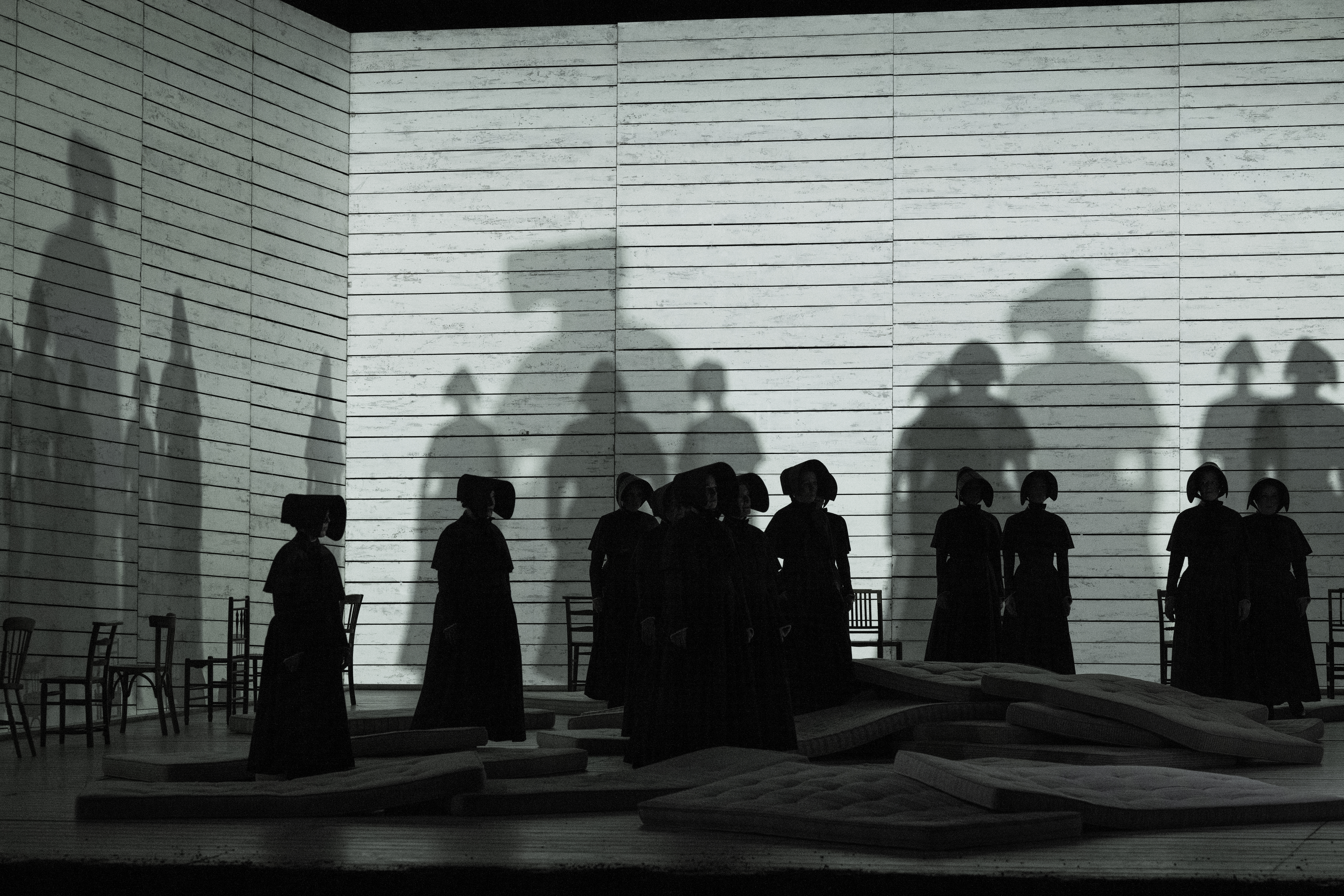 Women dressed in black stand in front of a white wall. Their shadows are projected onto the wall behind them.