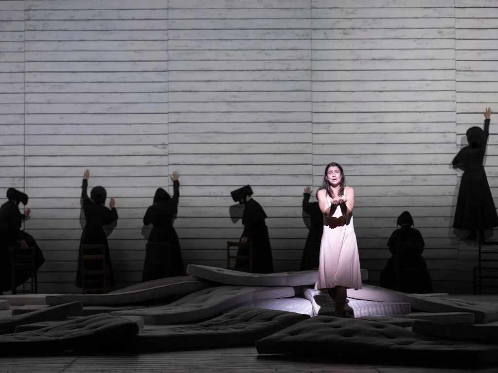 Several actors in black cloaks put their arms up to three white walls with their back to the audience. A woman in a white dress is in the centre of the stage.