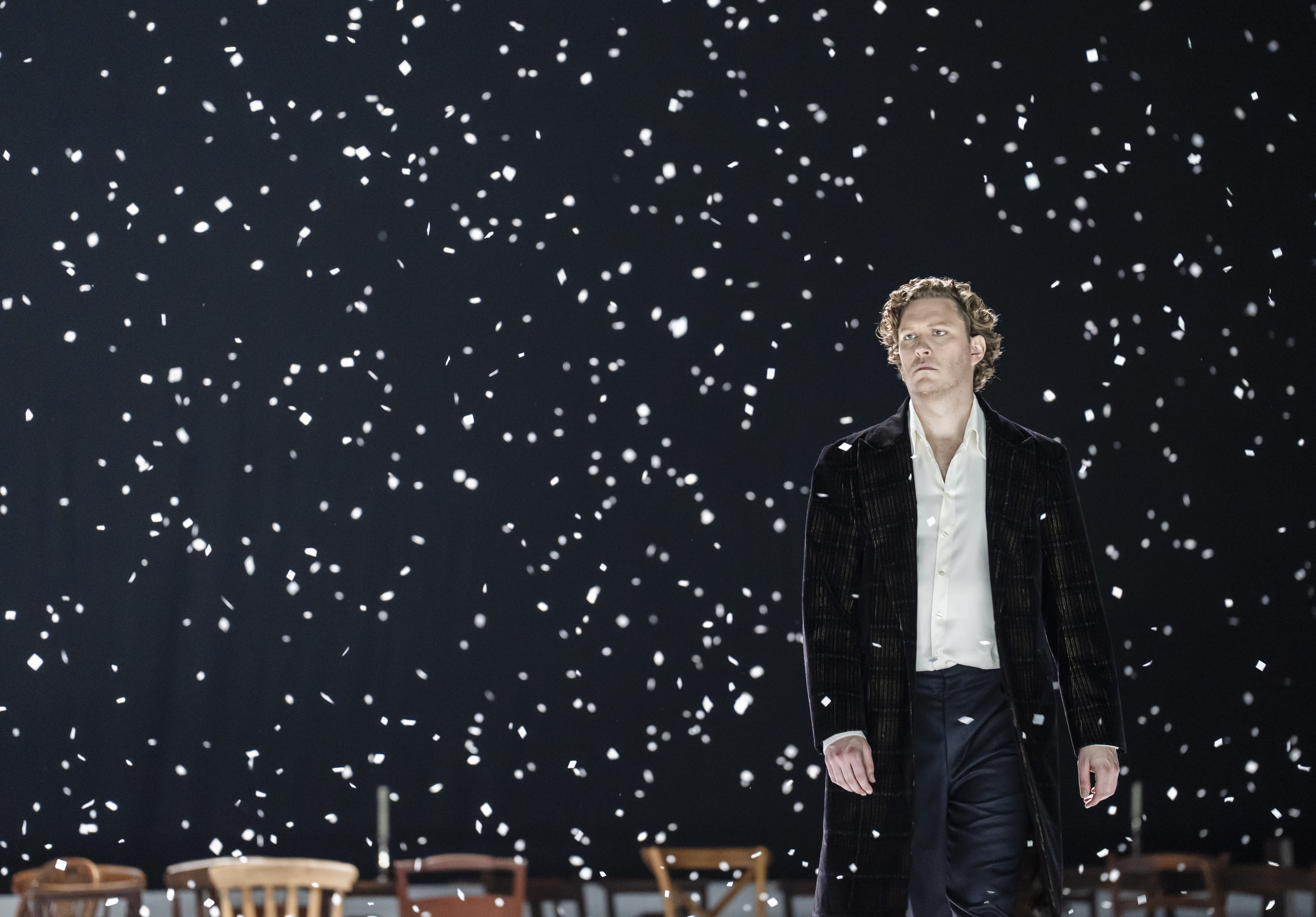 A man stands alone in the snow wearing a black suit jacket and white shirt. He looks out into the audience. 