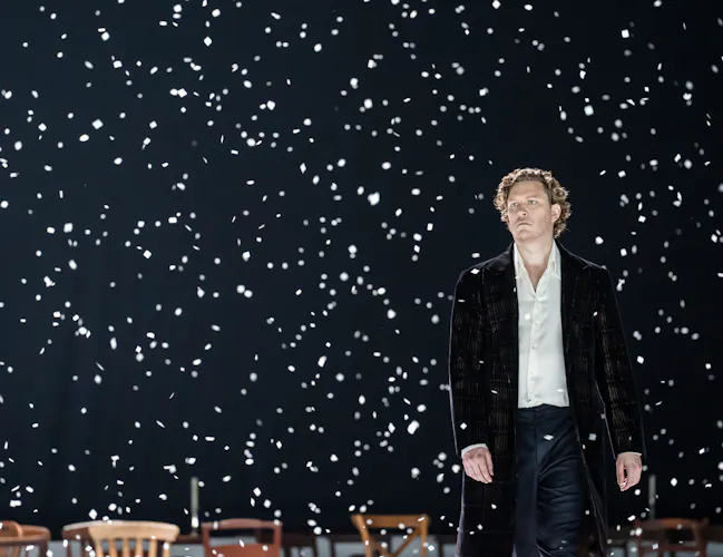 A man stands alone in the snow wearing a black suit jacket and white shirt. He looks out into the audience.