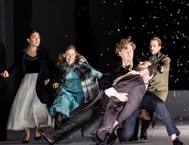 A man has been shot and falls into his friends arms. Behind them, their other friends run forwards. There is snow falling.