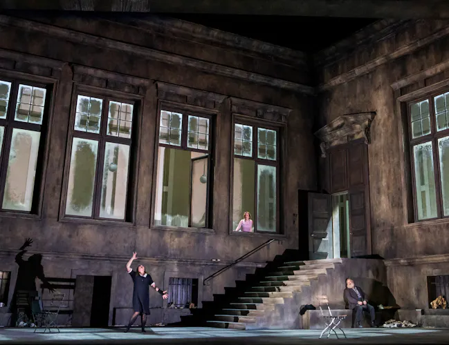 A large room with tall windows and a staircase set into the back corner. A woman in a purple leans slightly out of one of the windows watching as a woman in a black maid's dress screams, raising her hands upwards.