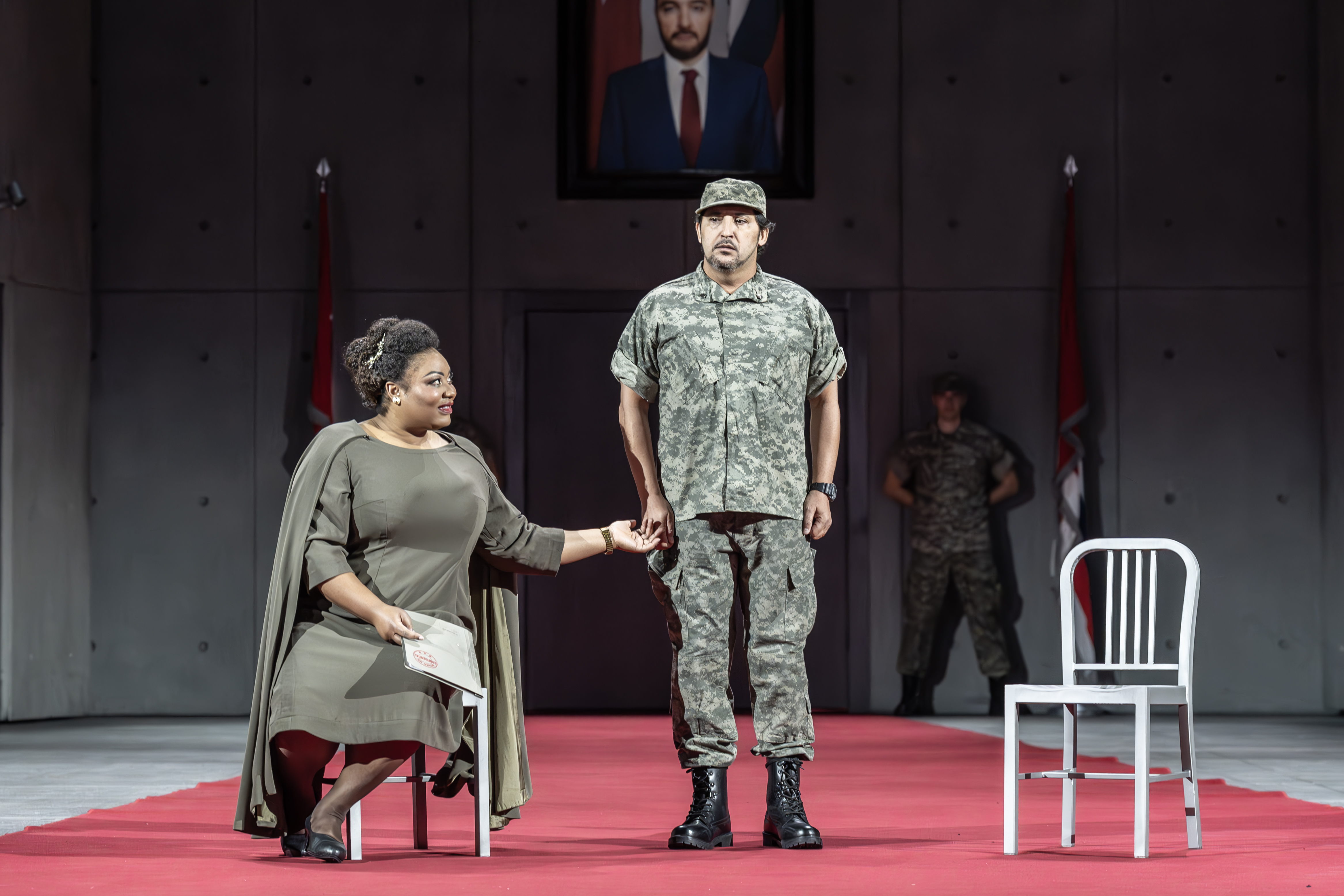 Amneris sits in one of two chairs onstage. She is wearing a military green dress. A soldier stands centre stage. 
