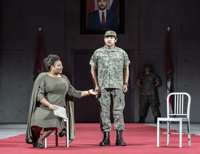 Amneris sits in one of two chairs onstage. She is wearing a military green dress. A soldier stands centre stage.