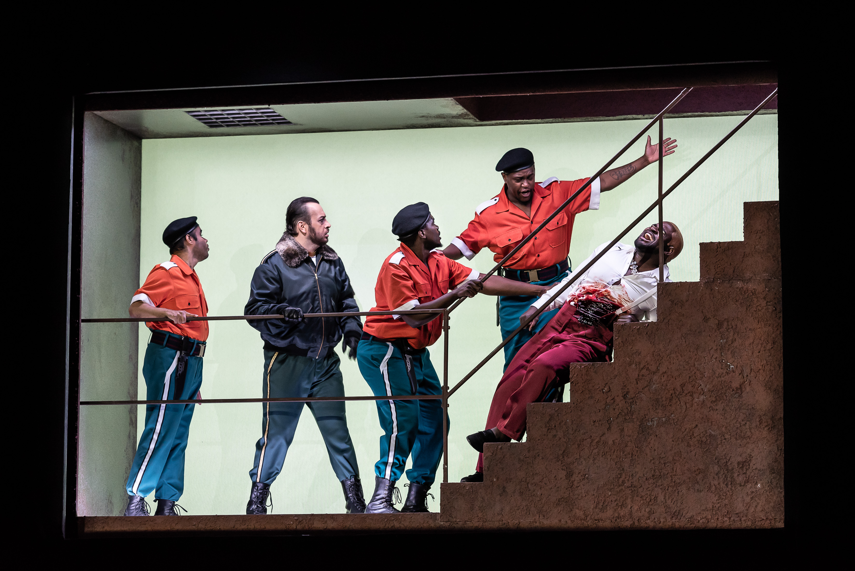 Four uniformed officers confront a wounded man leaning back on stairs, appearing distressed. One officer grabs the injured mans arm, while another points at him.