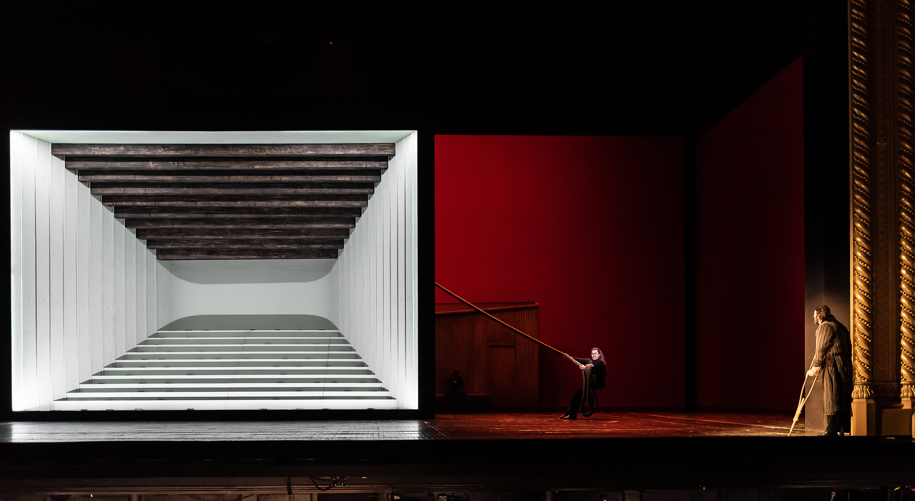 A theater stage with a modern set: a large white box with horizontal beams on the left and red walls on the right. Two performers stand apart, one holding a long pole extending toward the other.