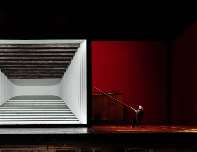 A theater stage with a modern set: a large white box with horizontal beams on the left and red walls on the right. Two performers stand apart, one holding a long pole extending toward the other.