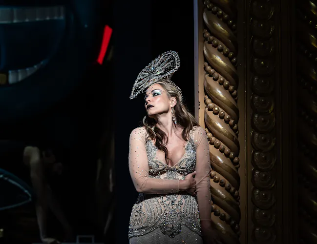 A woman in an ornate, sparkling dress and elaborate headpiece leans against a gold, decorative column on stage, looking thoughtful.