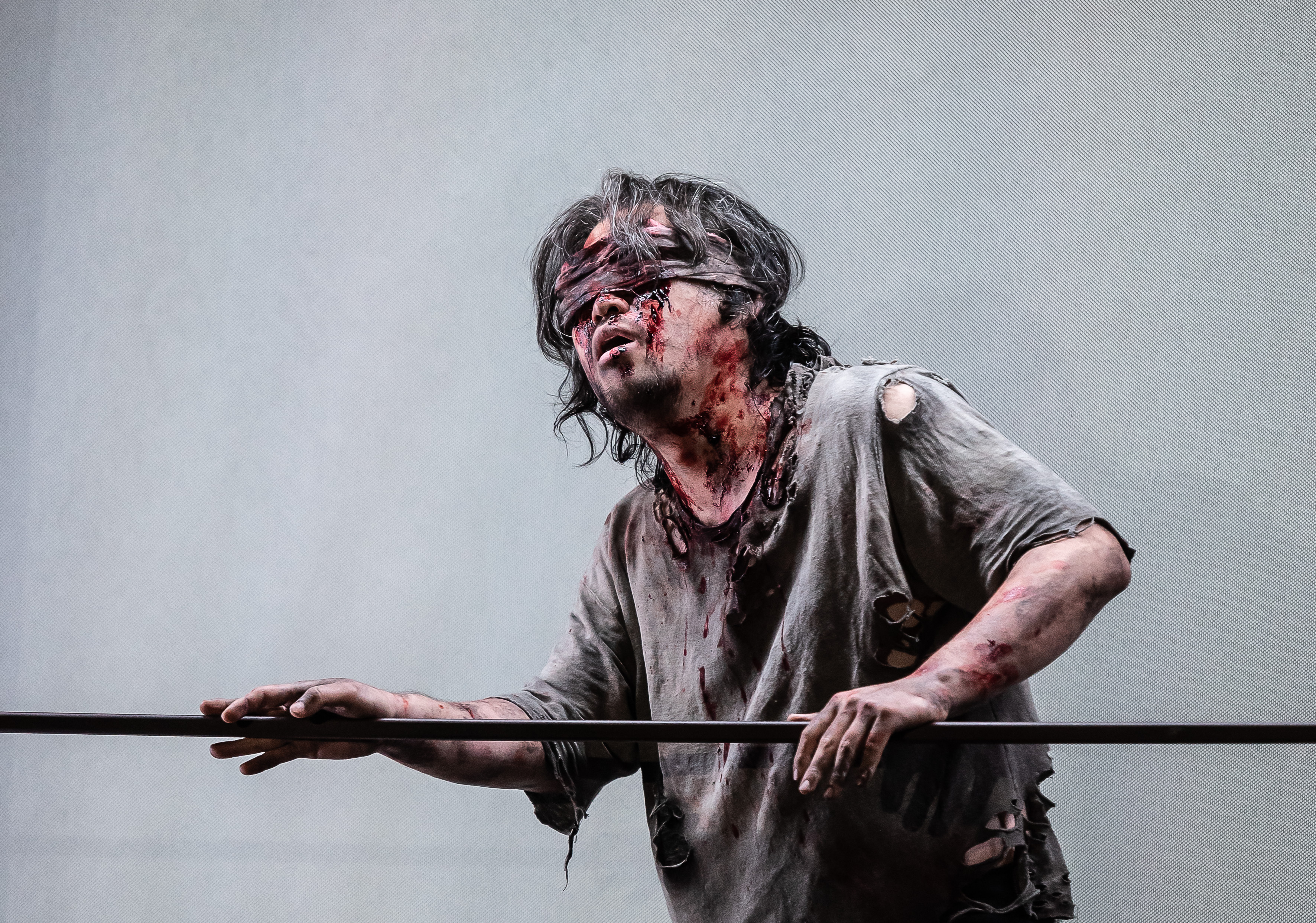 A blindfolded man covered in blood guides himself along a rope. 