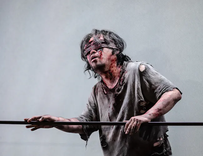 A blindfolded man covered in blood guides himself along a rope.