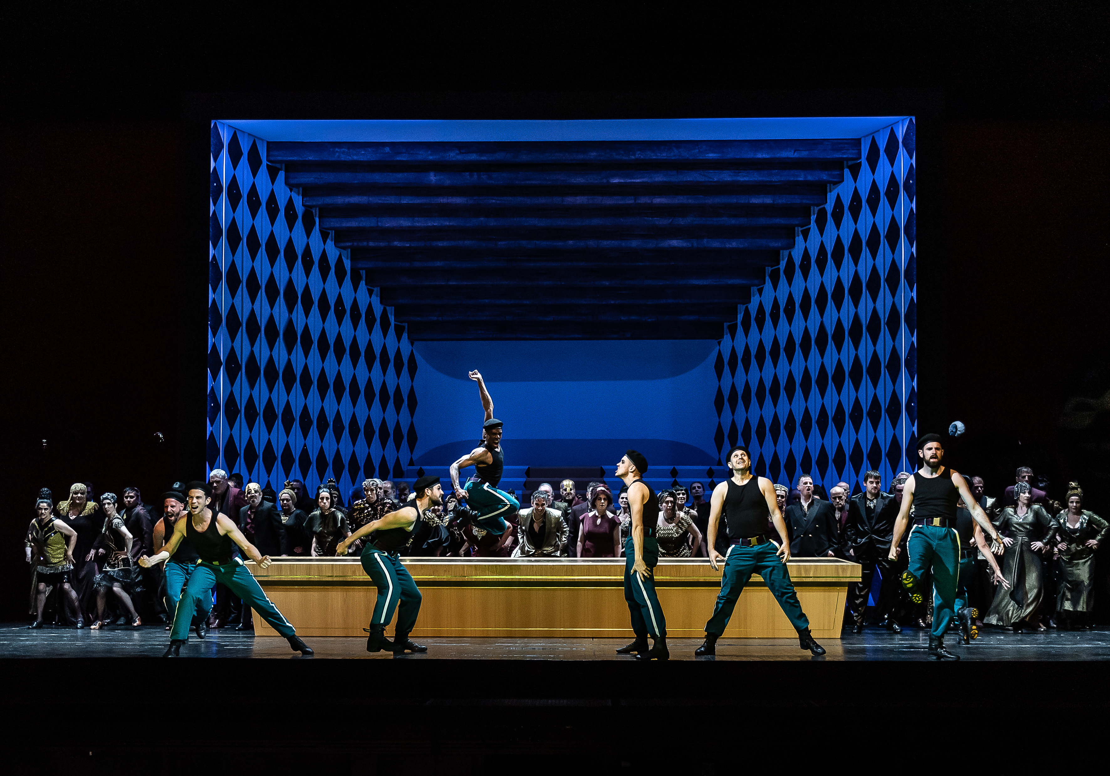 	 A group of dancers in green pants and black tops perform energetically at the front of a stage, with a cheering cast dressed in formal wear and a blue, diamond-patterned backdrop behind them.