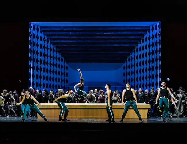 A group of dancers in green pants and black tops perform energetically at the front of a stage, with a cheering cast dressed in formal wear and a blue, diamond-patterned backdrop behind them.