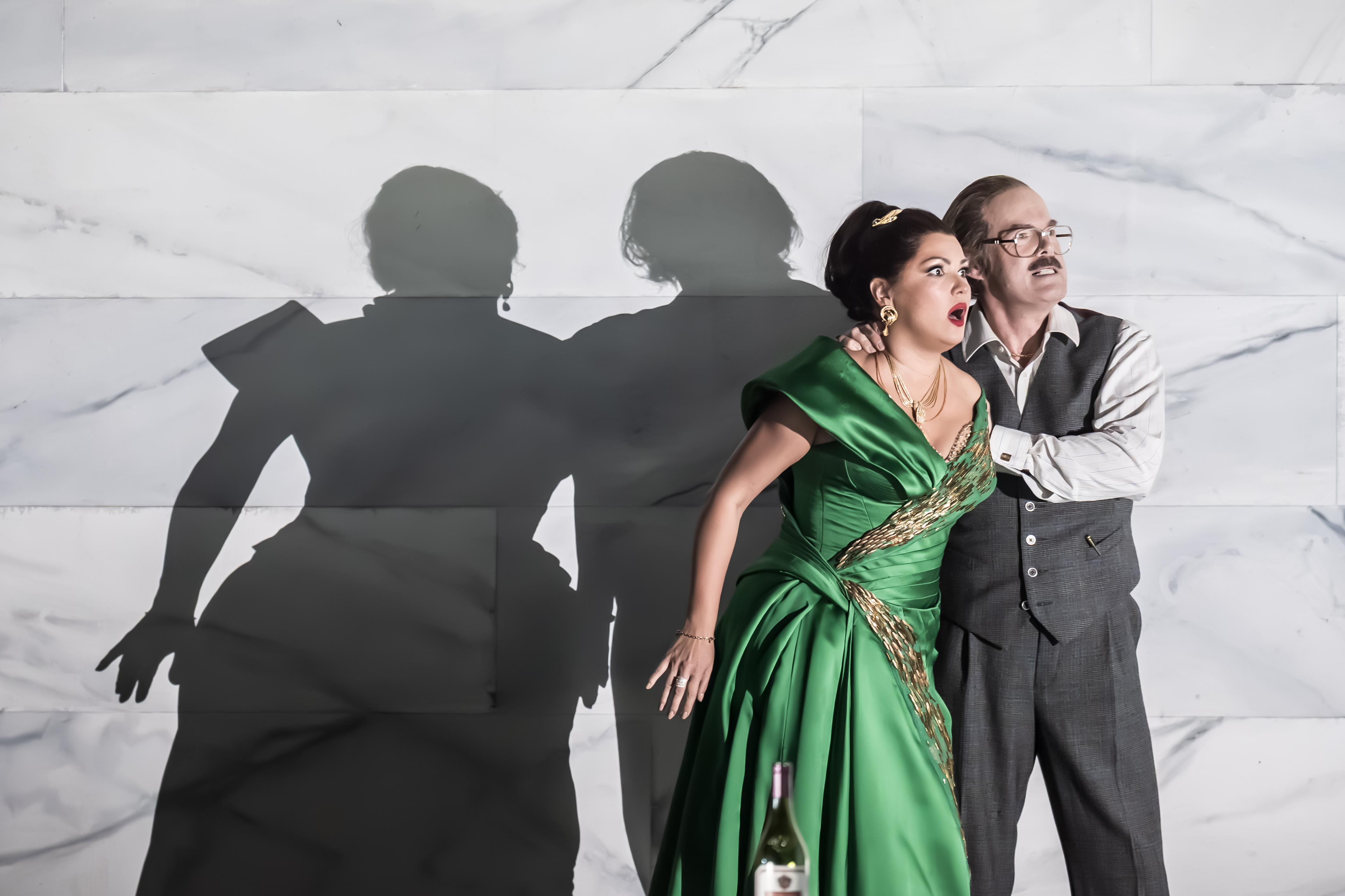 A woman in a green gown and a man in a vest stand close together against a marble wall, looking alarmed. Their dramatic shadows are cast on the wall behind them. A wine bottle sits in the foreground.