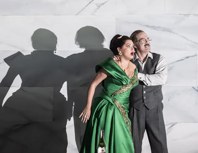 A woman in a green gown and a man in a vest stand close together against a marble wall, looking alarmed. Their dramatic shadows are cast on the wall behind them. A wine bottle sits in the foreground.