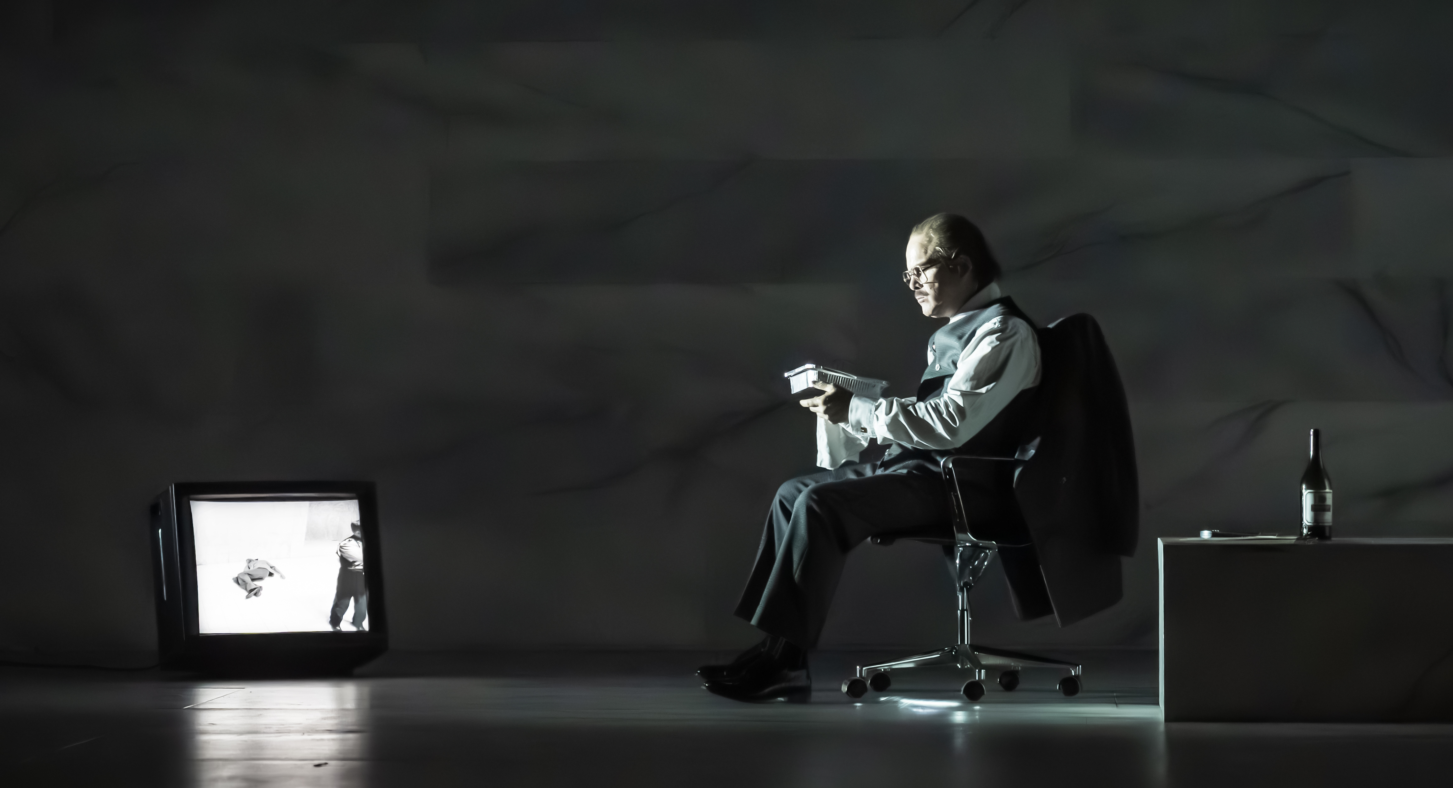 A man in formal attire sits on an office chair, reading a book under a dim light. A TV on the floor displays a black-and-white image, and a wine bottle sits on a low table nearby.