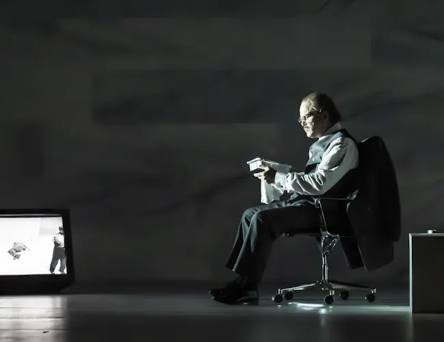 A man in formal attire sits on an office chair, reading a book under a dim light. A TV on the floor displays a black-and-white image, and a wine bottle sits on a low table nearby.