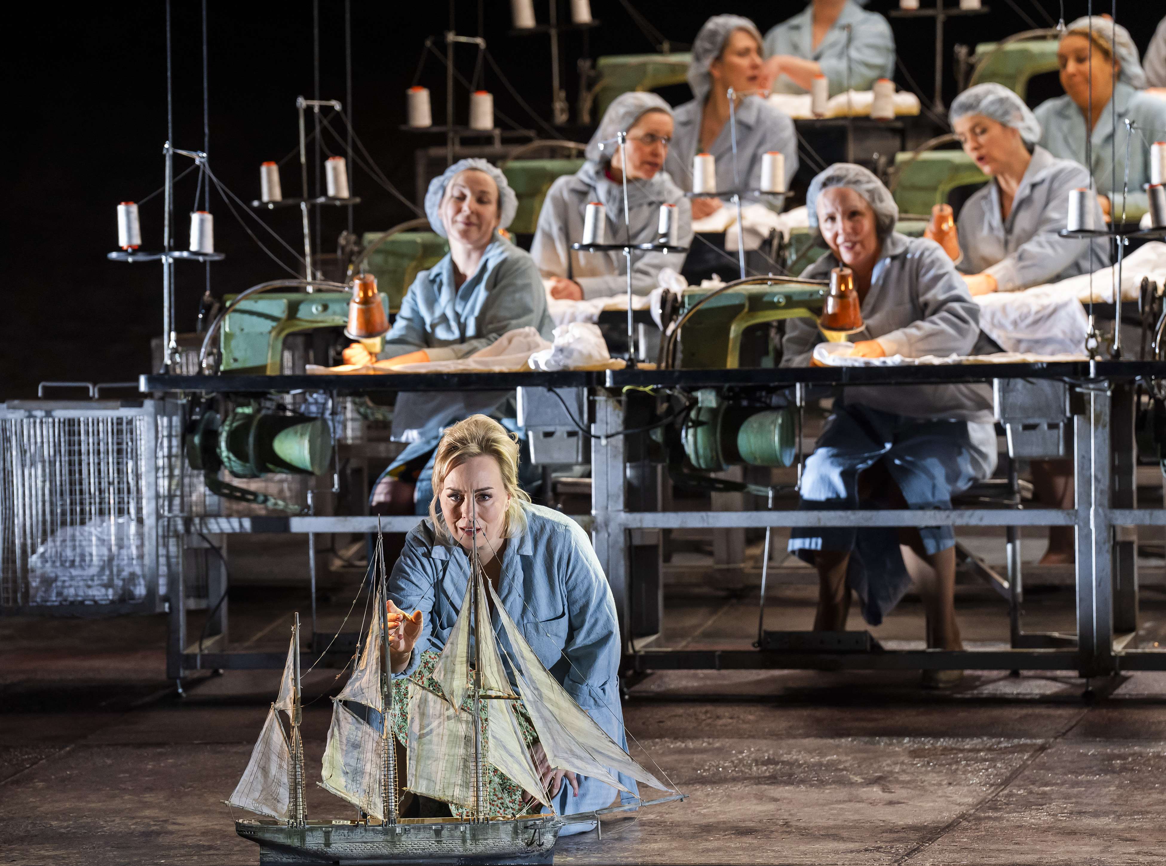 A woman kneels looking at a model ship. It had four sails. Behind her sit women at industrial sewing machines. 
