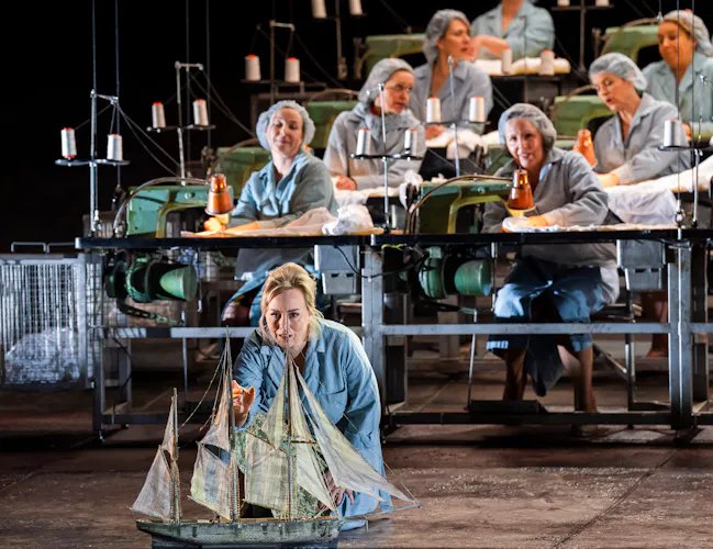 A woman kneels looking at a model ship. It had four sails. Behind her sit women at industrial sewing machines.